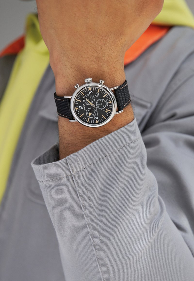 Black chronograph watch with silver accents, textured black strap, round dial, oversized numerals, and three sub-dials on a wrist.