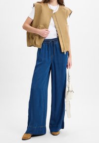 Beige gepolsterte Weste mit Reißverschluss, getragen über einem weißen T-Shirt, kombiniert mit lockeren blauen Jeans mit weitem Bein und braunen Schuhen.
