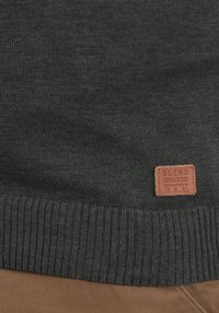 Mørkegrå strikket sweater med ribbet kant og et brunt lædretikette med "BLEND" prentet. Stoffet har en glat tekstur.