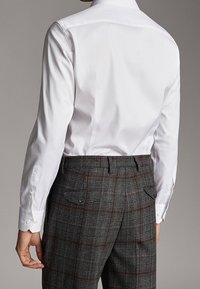 Man in een goed zittend wit shirt met lange mouwen, dat is gestopt in hooggewaande grijs geruite pantalon met knoopzakken aan de achterkant.