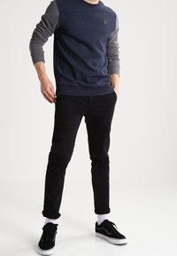Granatowy sweter crewneck z szarymi rękawami, czarne spodnie slim fit oraz czarne sneakersy z białymi podeszwami. Model pozuje na jasnym tle.