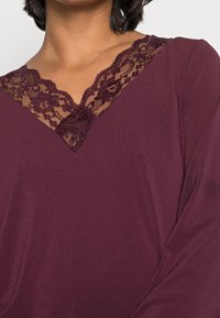 Burgundowa bluzka z dekoltem w serek, ozdobiona koronkowym wykończeniem przy dekolcie. Miękka tkanina o gładkiej strukturze i długimi rękawami.