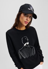 Hanorac negru cu o schiță albă a unei figuri pe față. Modelul poartă o șapcă neagră cu un design brodat.