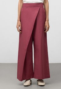 Wijde broek in een diepe rozenkleur, met een asymmetrisch wikkeldesign en een soepele textuur, gecombineerd met lichtbeige instappers.