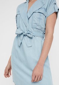 Ljusblå kortärmad klänning med slagkrage, bröstfickor och knytbälte i midjan, bärs av en person med avslappnade armar.