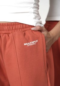Close-up af en person iført rustfarvede sweatpants med "MULTIPLY APPAREL" logo, hånden hvilende i forlommerne, parret med en hvid, rynket top.
