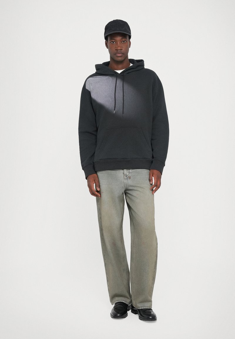 Schwarzer Kapuzenpullover mit grauem Verlaufsmuster, Kängurutasche; kombiniert mit hellgrauen geraden Jeans und schwarzen Schuhen. Schwarze Mütze.