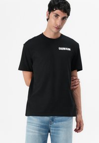 Juoda medvilnės marškinėliai trumpomis rankovėmis ir apvaliu kaklu, su baltu "CALVIN KLEIN" logotipu viršutiniame kairiajame krūtinės kampe.