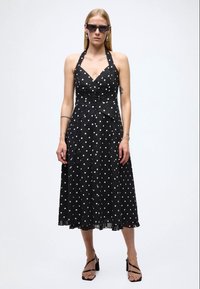 Vestido negro con lunares, con un corpiño ajustado, escote halter y falda fluida. La tela es ligera y de textura suave. Acentuado con joyería minimalista.