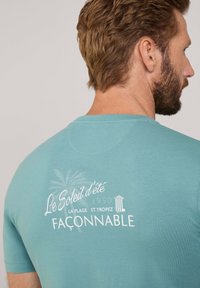 Camiseta de algodón turquesa con cuello redondo, mangas cortas y texto impreso en blanco con un diseño de palmera en la parte posterior.