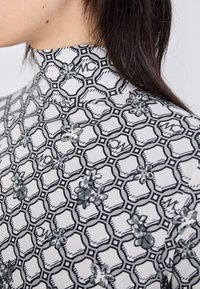 Durchsichtige Bluse mit hohem Kragen, die ein schwarz-weißes geometrisches Muster mit floralen Akzenten zeigt, aus leichtem, strukturiertem Stoff gefertigt.