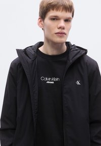 Μαύρο ζακέτα με φερμουάρ και υφή, με κουκούλα. Κάτω από αυτή, μαύρο μπλουζάκι με λευκό λογότυπο "Calvin Klein JEANS".