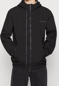 Svart vindjacka med dragkedja fram, elastiska ärmslut och huva. Har reflexdetaljer och logotyp på bröstet och sidan.