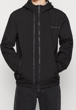 Schwarze Windjacke mit Reißverschluss, elastischen Bündchen und Kapuze. Verfügt über reflektierende Akzente und ein Logodetail auf der Brust und der Seite.