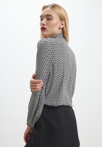 Blusa a pois bianco e nero con un design geometrico a foglia, maniche lunghe a sbuffo e colletto alto, abbinata a una gonna nera.