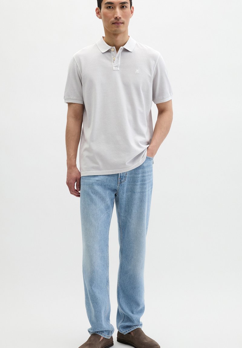 Mann steht und trägt ein hellgraues Poloshirt, hellblaue Jeans und braune Schuhe vor einem schlichten weißen Hintergrund.