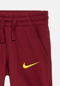 Calças de fato de treino em bordeaux com um tecido de algodão texturizado. Apresenta um logotipo amarelo da Nike na frente, cintura elástica e bolsos laterais.