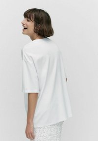 Vit oversized t-shirt med korta ärmar, slät textur, rund halsringning och avslappnad passform, bärs med en strukturerad vit kjol.