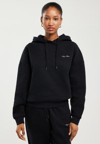 Naine, kes kannab musta kapuutsiga pusa ja sobivaid musti pükse, millel on väikene valge märk rinnal ja reiel, seisab ühetaolise tausta ees.