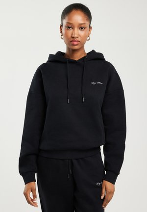 Kobieta w czarnej bluzie z kapturem oraz dopasowanych czarnych spodniach z małym białym logo na piersi i udzie, stojąca na jednolitym tle.