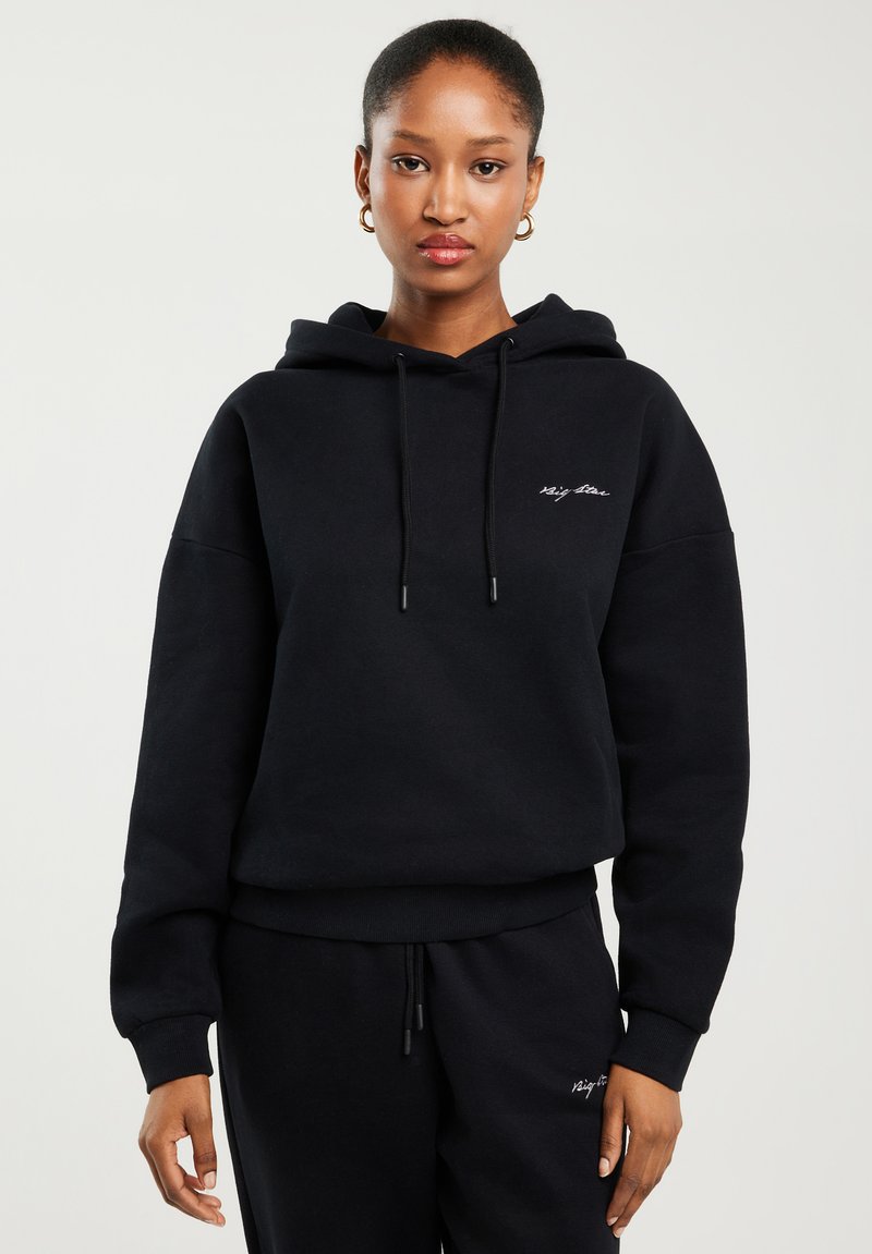 Naine, kes kannab musta kapuutsiga pusa ja sobivaid musti pükse, millel on väikene valge märk rinnal ja reiel, seisab ühetaolise tausta ees.