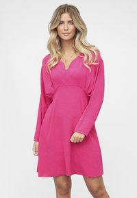 Vaaleanpunainen mekko, jossa on pitkät hihat, V-aukko ja rento istuvuus. Valmistettu pehmeästä, sileästä kankaasta, jossa on hieman teksturoitu pinta.