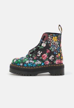 Bota negra con suela gruesa y cordones, con estampado floral multicolor y costuras amarillas a lo largo del borde de la suela, vista lateral sobre fondo blanco.