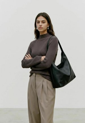 Femme en pull côtelé marron à col montant et pantalon beige plissé, debout les bras croisés, portant un sac à main en cuir noir.