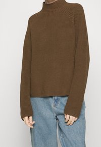 Person som bär en brun ribbad stickad tröja med långa ärmar och ljusblå denimjeans, stående mot en enkel vit bakgrund.