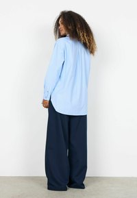 Sieviete ar cirtainiem matiem stāv sāniski, valkājot gaiši zilu oversized kreklu un plašām tumši zilaņģēm biksēm.