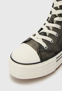 Sneaker montante en tissu gris avec bout en caoutchouc blanc, présentant des lacets blancs, des œillets en métal et une semelle en caoutchouc rayée.