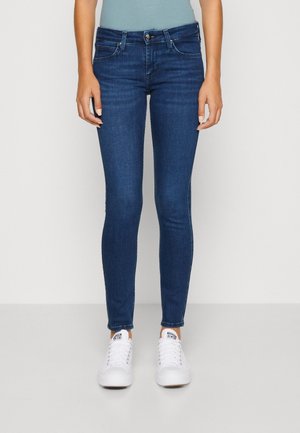 Jeans de corte ajustado de color azul oscuro, hechos de denim, con un clásico diseño de cinco bolsillos y sutiles detalles de costura, combinados con zapatillas blancas.