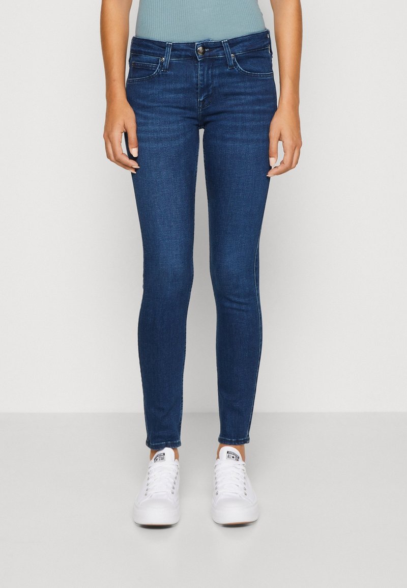 Jeans de corte ajustado de color azul oscuro, hechos de denim, con un clásico diseño de cinco bolsillos y sutiles detalles de costura, combinados con zapatillas blancas.