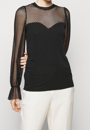 Donna che indossa un top nero con maniche lunghe trasparenti e motivo, e pannello sul petto, abbinato a pantaloni bianchi, in piedi davanti a uno sfondo semplice.