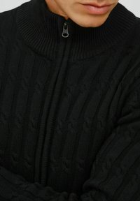 Pull en tricot noir avec un col haut, une fermeture éclair à l'avant et des motifs en relief pour ajouter de la texture. Vue en gros plan mettant en avant les détails du tricot.