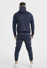 Navyblauwe hoodie met een hoge kraag en zwarte manchetten, gecombineerd met bijpassende joggers. Soepele stof met een slank ontwerp en elastische tailleband.