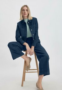 Donker spijkerjack met knoopsluiting en twee voorkruiszakken, gecombineerd met bijpassende wijde broek. Lichtgroene top eronder en beige instappers.