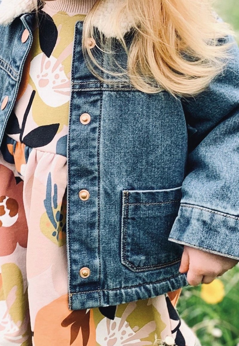 Enfant portant une robe fleurie et une veste en jean bleue avec col en laine, debout dehors sur de l'herbe avec des fleurs jaunes.