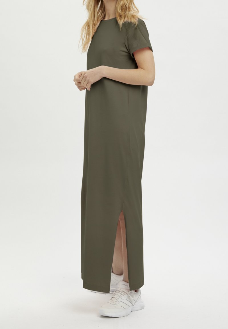 Femme portant une robe longue olive à manches courtes avec une fente latérale, associée à des chaussures de sport blanches, debout sur un fond blanc.