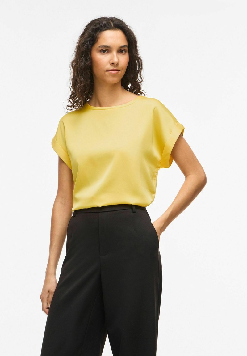 Femme aux cheveux bouclés portant un chemisier jaune à manches courtes rentré dans un pantalon noir taille haute, debout devant un fond uni.