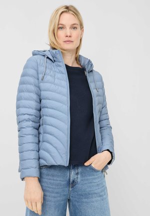 Frau trägt hellblaue gesteppte Kapuzenjacke, marineblauen Pullover und blaue Jeans, steht mit einer Hand in der Tasche vor einfarbigem Hintergrund.