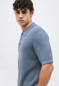 Νέος άνδρας με σγουρά σκούρα μαλλιά, που φοράει μπλε πόλο μπλουζάκι με υφή και κοντά μανίκια, κοιτάζει ελαφρώς πλάγια σε απλό φόντο.