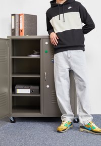 Metallskåp i grått med hyllor; vita och gröna runda knopp. Person iklädd svart huvtröja, ljusa jeans och flerfärgade sneakers.