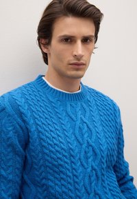 Blauer Strickpullover mit Zopfmuster, geripptem Rundhalsausschnitt, strukturierten Mustern und lockerem Schnitt, kombiniert mit einem weißen Unterhemd.