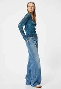 Teal doorzichtige top met lange mouwen en tekstpatronen, gecombineerd met lichtblauwe wijde denim jeans. De outfit heeft een relaxte pasvorm en subtiele gerimpelde randen.