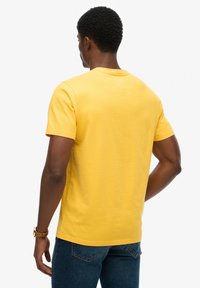 Kortærmet, rundhalset gul bomuldst-shirt vist bagfra, med en glat tekstur og en afslappet pasform. Brugt sammen med blå jeans.