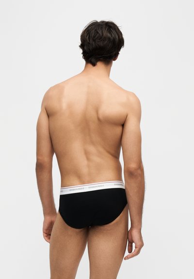 Homme debout, dos tourné, portant un slip noir avec une ceinture blanche de la marque "DSQUARED2", sur un fond clair uni.