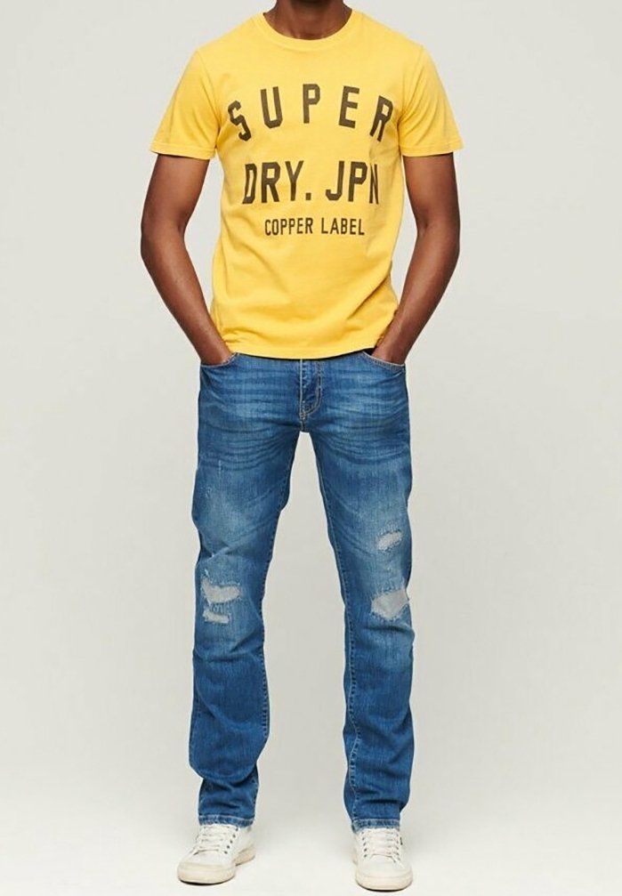 Gul bomulds t-shirt med sort trykt tekst, parret med blå slidte denim jeans og hvide sneakers. Simplet, afslappet outfit.