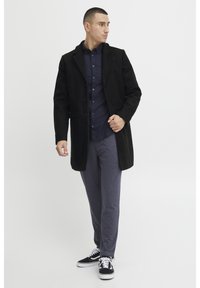 Svart ullblandingsfrakk med spiss krage og frontknapper, kombinert med en mørk skjorte med knapper og grå joggebukser, fullført med svarte sneakers.