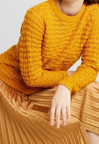Pletený žlutý svetr s texturovaným vzorem, spárovaný s plisovanou zlatou saténovou sukní s knoflíkovými detaily po straně.